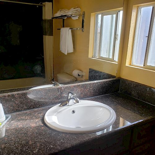 A bathroom sink with a rounded white basin, a granite countertop, a mirror, soap and cup holder, towel rack, toilet, window, and wall-mounted shelves.