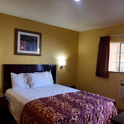 A cozy hotel room with a queen-sized bed, framed artwork, bedside lamps, a window with brown curtains, and an air conditioning unit.