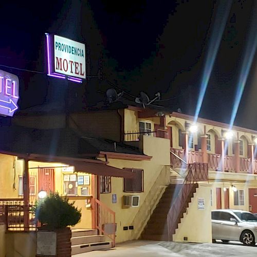 A brightly lit motel at night with neon signs and parked cars outside. The motel has two floors with exterior staircases.