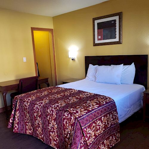 A hotel room with a bed, two nightstands, wall art, wall-mounted lights, and a desk with a chair; walls are yellow.