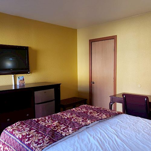 The image shows a hotel room with a bed, a TV mounted on the wall, a dresser, a mini-fridge, a desk with a chair, and a door.