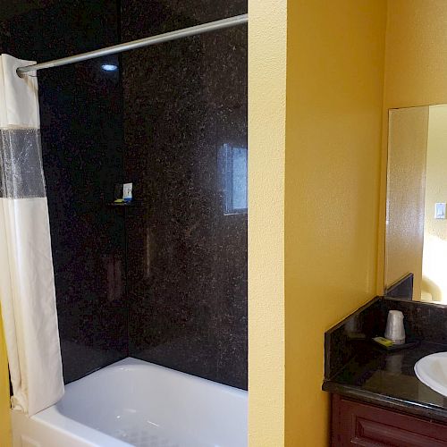 The image shows a bathroom featuring a bathtub with a shower curtain, a mirror above a sink, and yellow walls. There is a bed visible in the background.