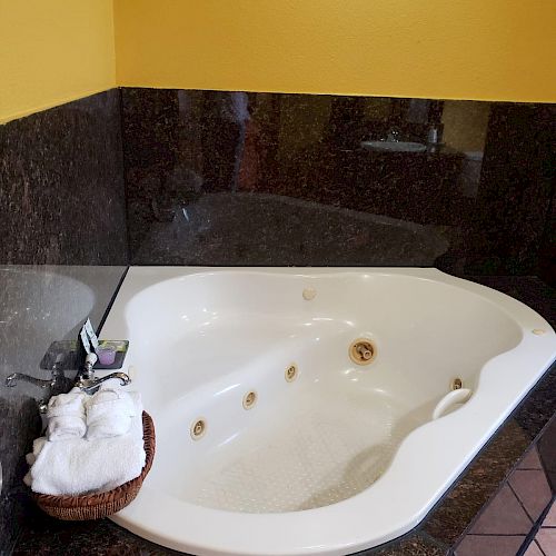 The image shows a corner bathtub with jets, surrounded by dark tiles. There are folded towels and a basket placed on the tub's edge.