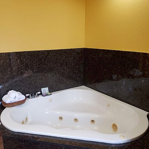 The image shows a white corner bathtub with jets, set against dark tiling. There is a rolled towel and some toiletries placed on the tub edge.