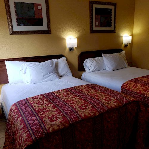 This image shows a hotel room with two neatly made beds, white linens, red patterned bedspreads, wall art, and two wall-mounted lamps.