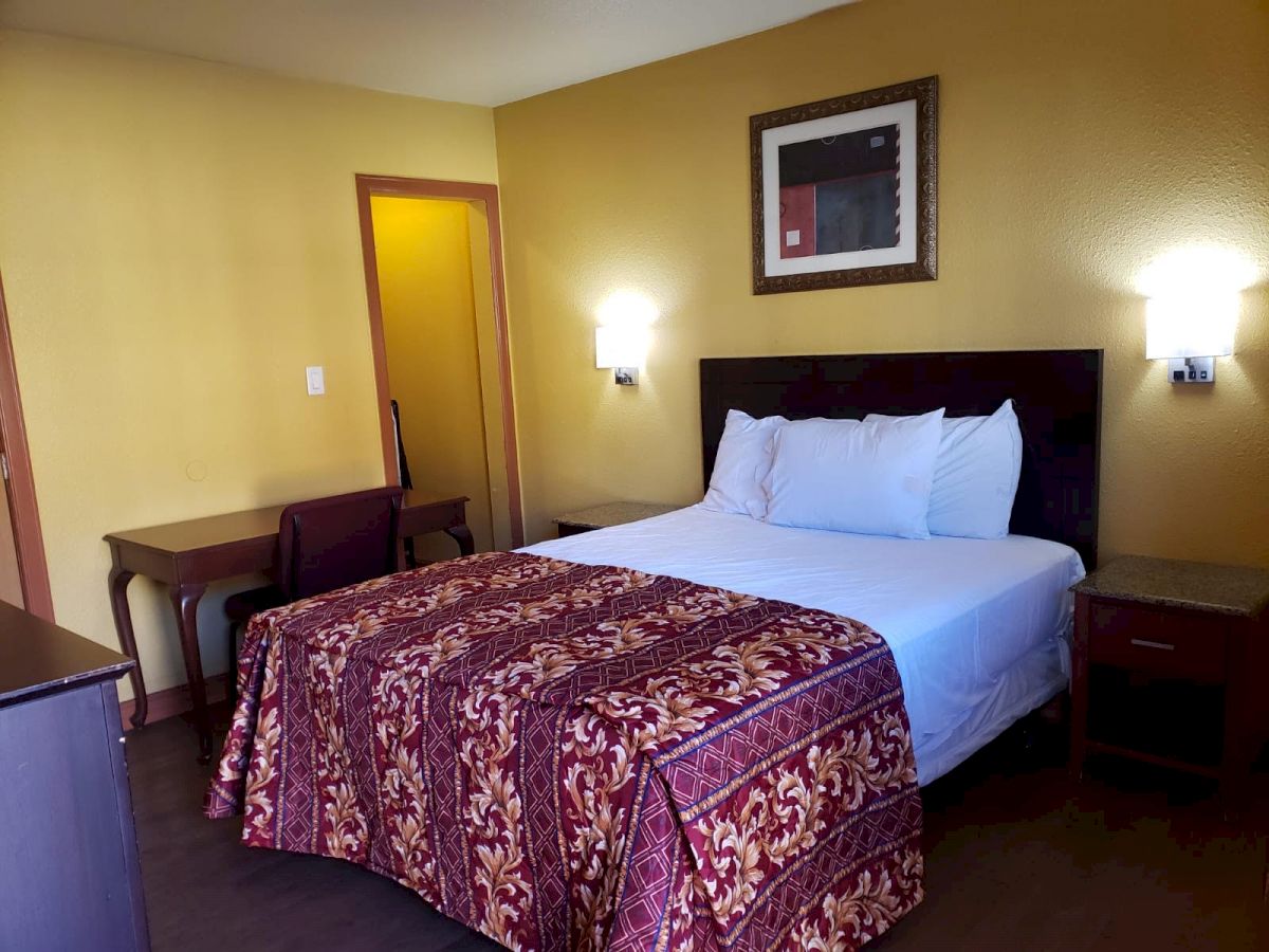 The image shows a neatly made bed with a colorful bedspread, flanked by two nightstands in a small, warmly lit room with a desk and chair in the corner.