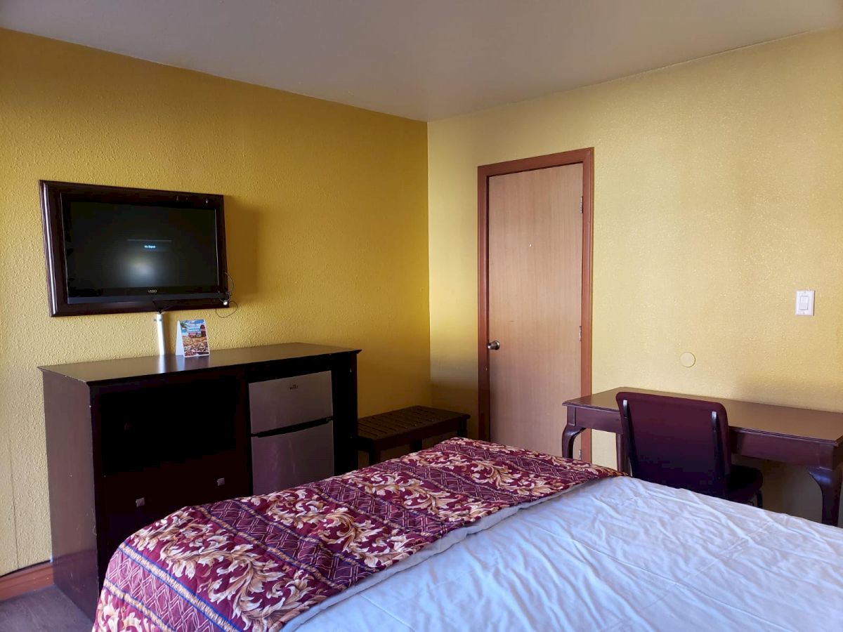 The image shows a cozy room with a bed featuring a red and white patterned blanket, a TV mounted on the wall, a desk, chair, and a door.