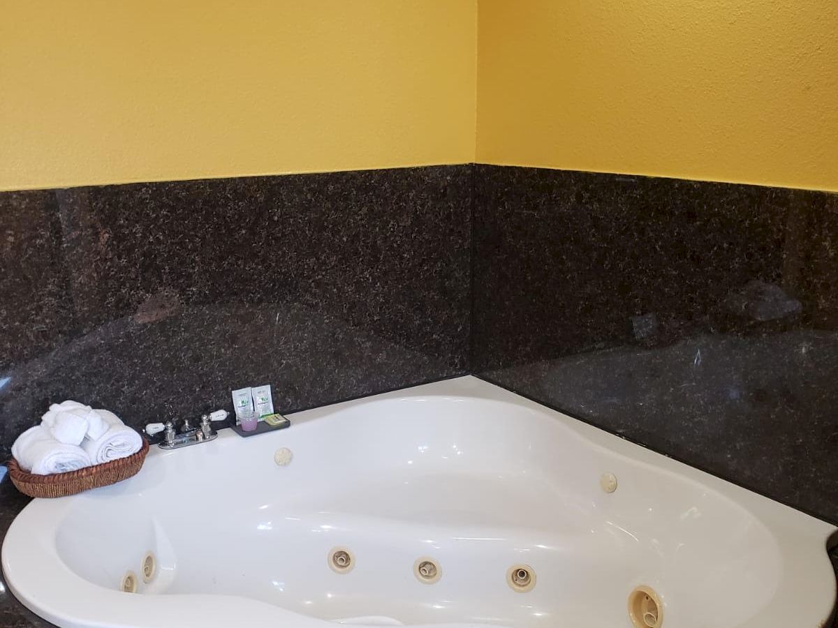 This image shows a corner jetted bathtub with dark tile surround and yellow walls, accompanied by a small basket holding towels and bath products.