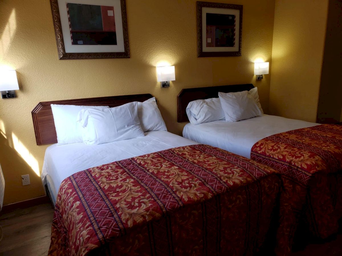 The image shows a hotel room with two neatly made beds, white linens, red and gold bed covers, wall lamps, and framed art above the beds.