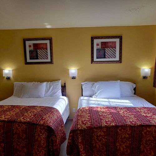 The image shows a hotel room with two neatly made beds, adorned with red patterned covers, and framed art on a yellow wall.