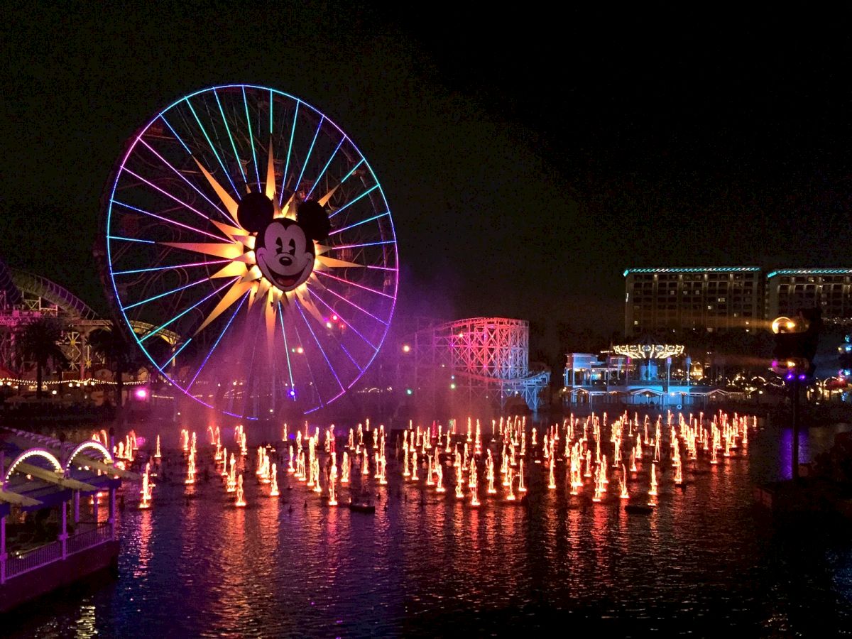 A night scene featuring a large illuminated Ferris wheel with a cartoon character face and a water fountain show with lights and buildings in the background.