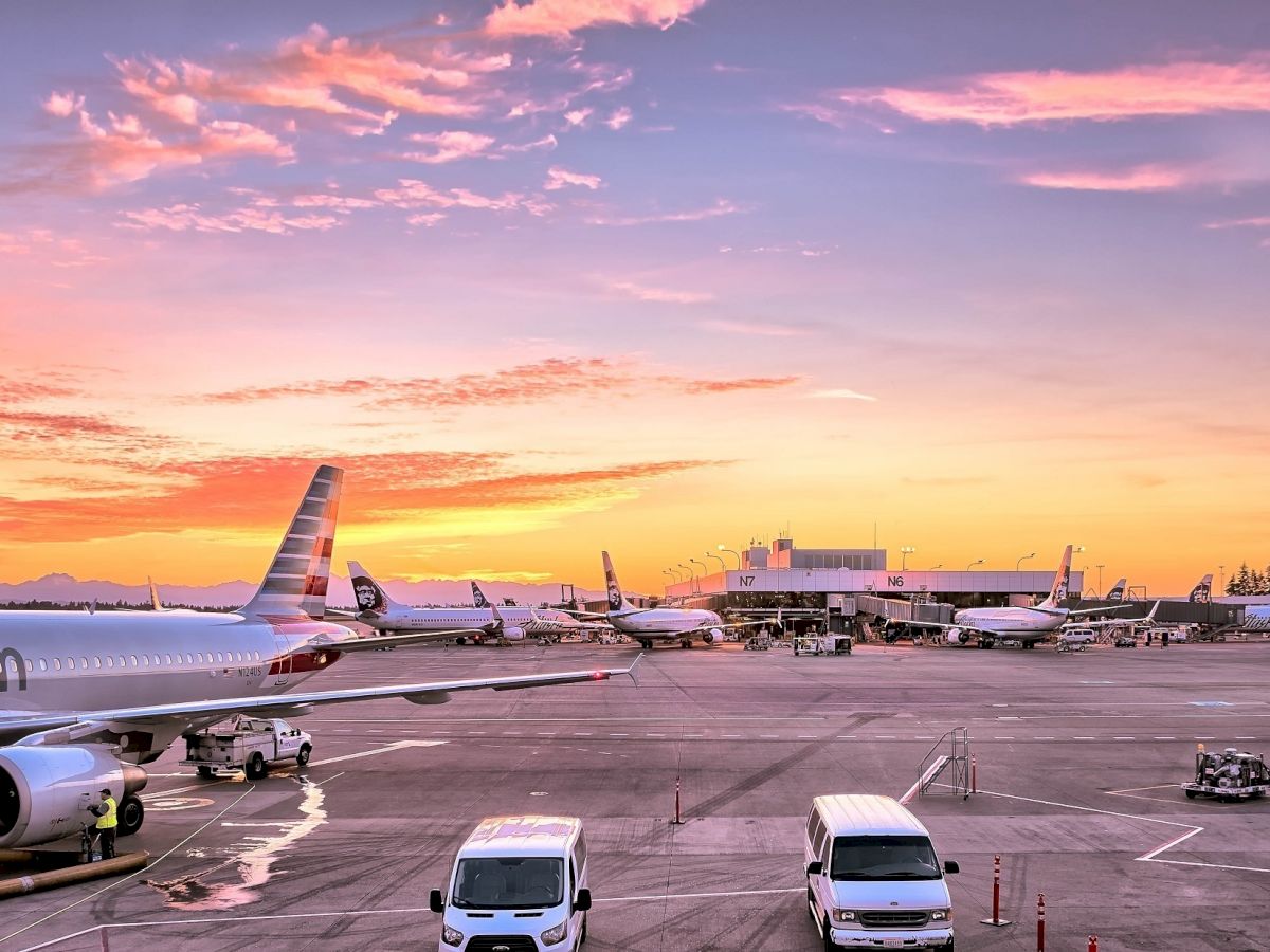 The image shows an airport tarmac at sunset with several airplanes, airport ground vehicles, and the terminal building in the background.