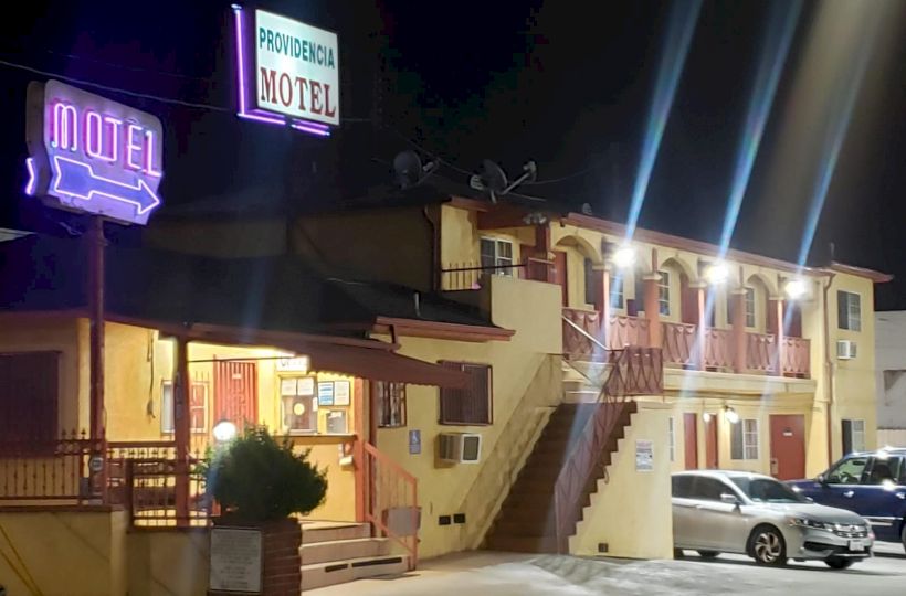 The image shows a two-story motel at night with neon signs and lights; some cars are parked in front, and stairs lead to the second floor.