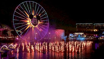 The image shows a large Ferris wheel with a Mickey Mouse face, colorful lights, and a fountain show at night in an amusement park.