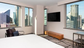 A modern hotel room with large windows showing a cityscape, a wall-mounted TV, a leather chair, and a bed in the foreground ending the sentence.