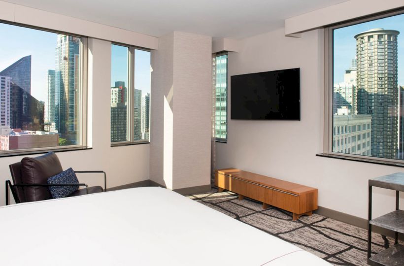 A modern hotel room with large windows showing a cityscape, a wall-mounted TV, a leather chair, and a bed in the foreground ending the sentence.