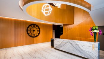 A modern lobby with a marble reception desk, wooden accents, a geometric chandelier, a wall clock, and a flower arrangement on the counter.