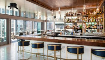 The image shows a modern bar with a wooden counter, blue and gold bar stools, a well-stocked liquor shelf, and large windows overlooking a city.