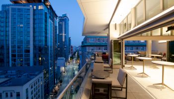 The image shows a modern balcony with seating and tables, overlooking a city street lined with tall buildings during the evening.