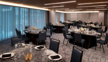 A banquet or conference room with round tables set for a formal event, featuring neatly arranged place settings, black napkins, and floral centerpieces.