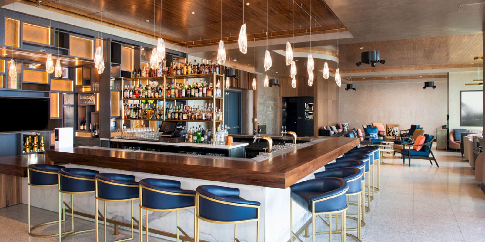 This image shows a modern bar with a wooden counter, blue bar stools, hanging lights, and a well-stocked liquor display.