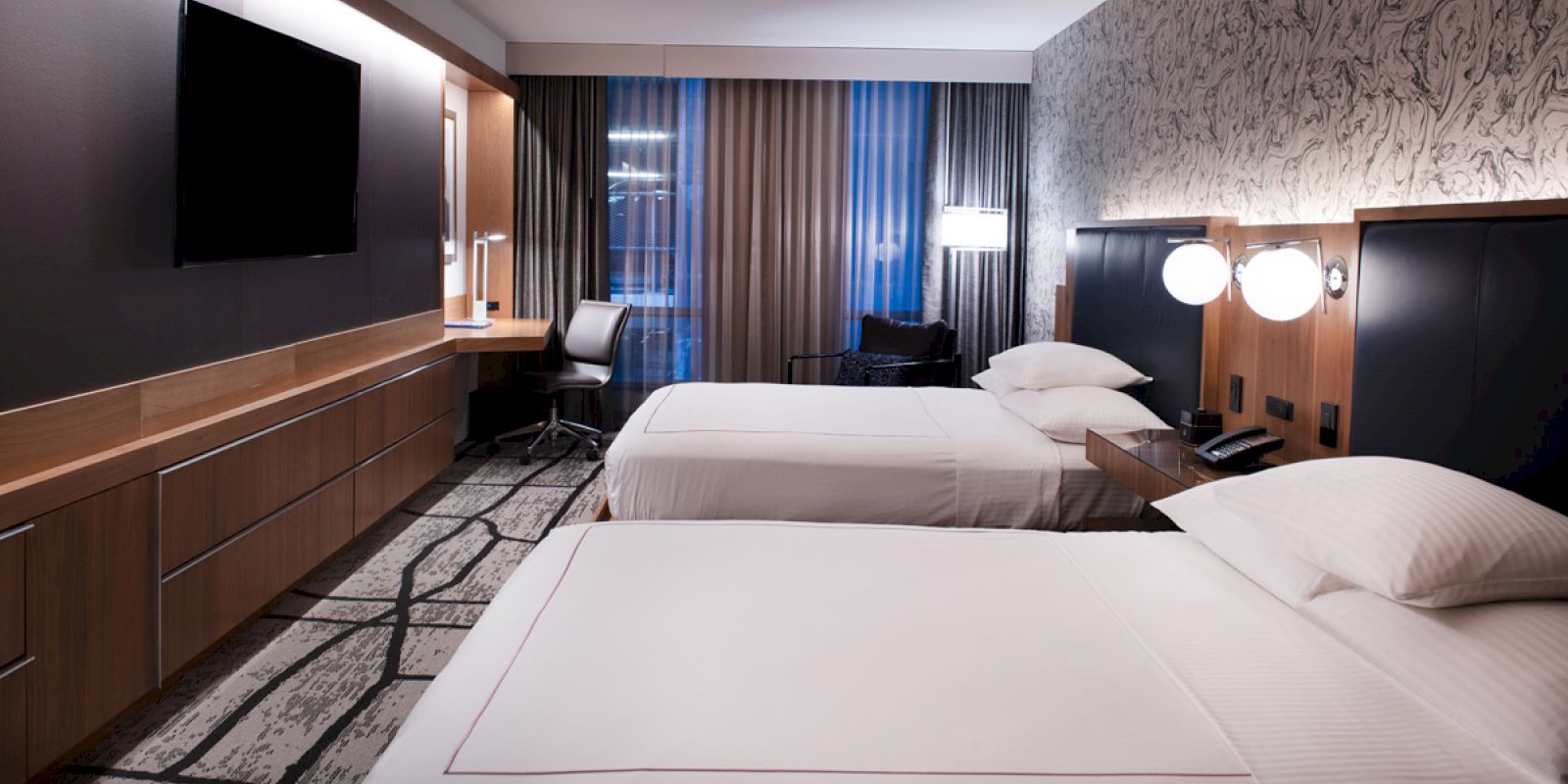 A modern hotel room with two beds, a desk, and a flat-screen TV on the wall. The room features stylish decor and lighting, with large windows.
