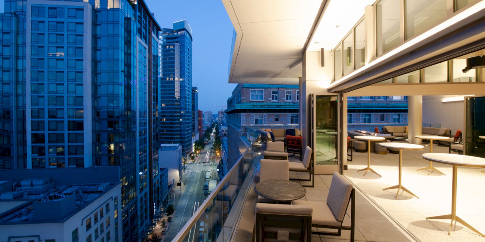 A modern balcony with tables and chairs overlooks a cityscape with high-rise buildings under a twilight sky.