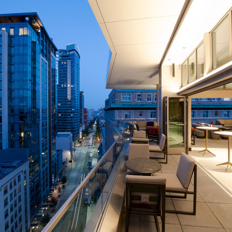 The image shows a modern cityscape view from a high-rise balcony at dusk, featuring outdoor seating areas and the interior of a contemporary building.