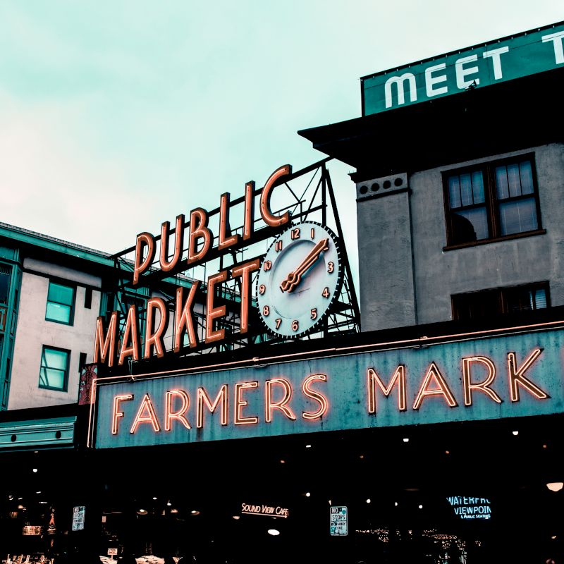 The image shows a public market with large neon signs reading "Public Market" and "Farmers Market" against a backdrop of older buildings and a cloudy sky.