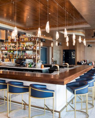 This image shows a modern bar area with hanging lights, a fully stocked bar, blue bar stools, and a lounge area with chairs in the background.