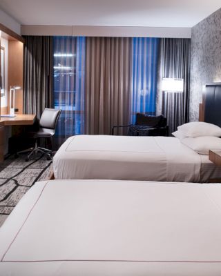 The image shows a modern hotel room with two neatly made beds, a wall-mounted TV, a desk with a chair, and a patterned carpet and wallpaper.