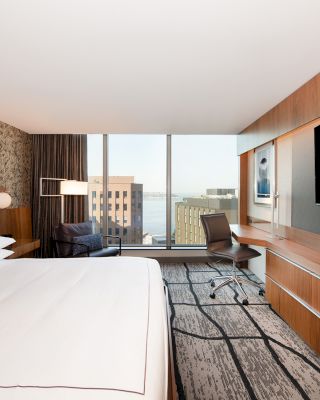 A modern hotel room with a large bed, wall-mounted TV, desk, chair, and a window with a city view. The decor is sleek and contemporary.