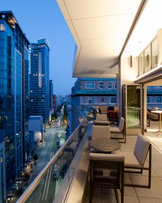 The image shows a modern urban balcony at dusk, overlooking a city street with tall buildings. The balcony has tables and chairs for seating.