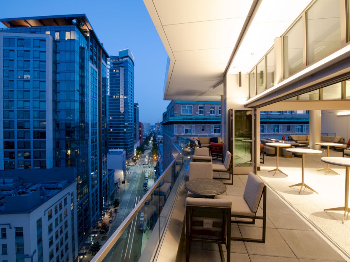 The image shows a rooftop terrace with tables and chairs, overlooking a cityscape at dusk, with tall buildings and a busy street below.