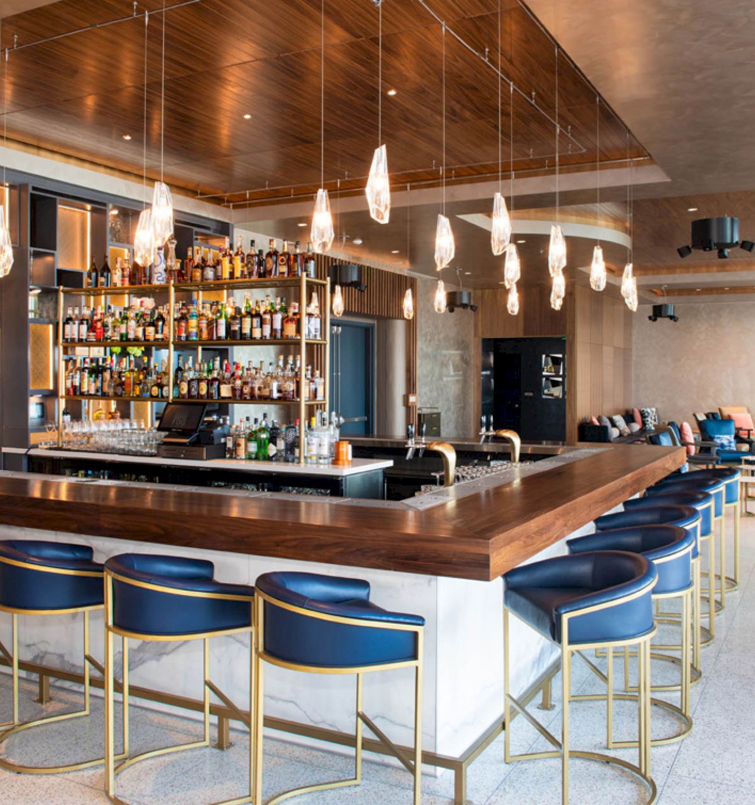 A modern bar with a wooden counter, blue and gold bar stools, a well-stocked liquor shelf, and hanging lights in a spacious, elegant room.