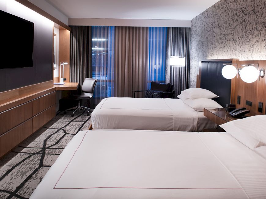 The image shows a modern hotel room with two neatly made beds, a large TV, a work desk near the window, and stylish decor and lighting.
