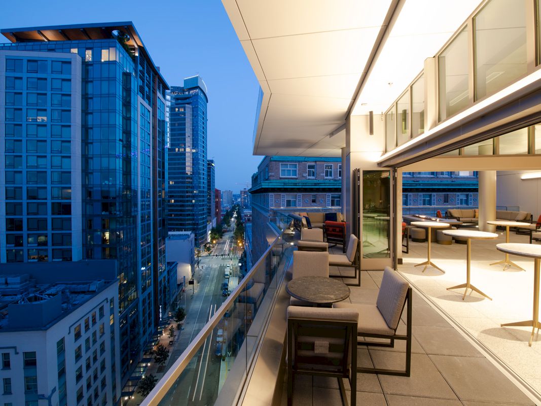 A modern balcony with chairs and tables overlooks a city street lined with tall, glass-covered buildings during dusk.