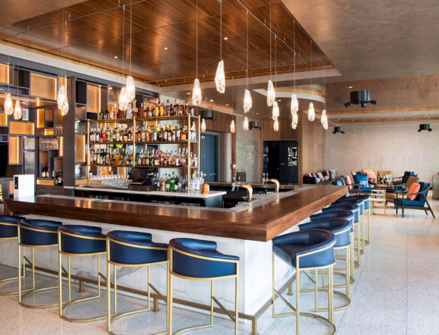 This image shows a modern bar with hanging lights, a well-stocked bar, and blue barstools. There are tables and chairs in the background.