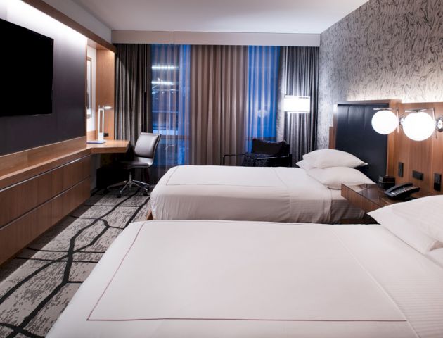 The image shows a modern hotel room with two neatly made beds, a desk, a chair, a wall-mounted TV, and stylish lighting and decor.