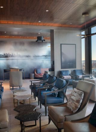 A modern lounge with armchairs, tables, large windows with a city view, and a mural of a city skyline on the wall, providing a cozy atmosphere.