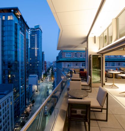 The image shows a modern cityscape with tall buildings and a well-lit balcony featuring tables and chairs in the foreground.
