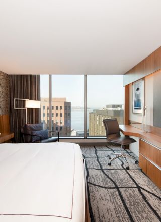 A modern hotel room with a large bed, a wall-mounted TV, a work desk, and a window offering a city and water view in the background.