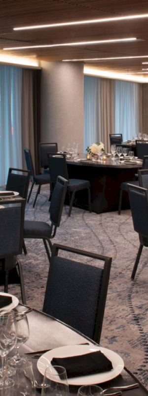 A beautifully lit banquet hall with round tables set for an event, featuring elegant table settings, black chairs, and light blue curtains in the background.