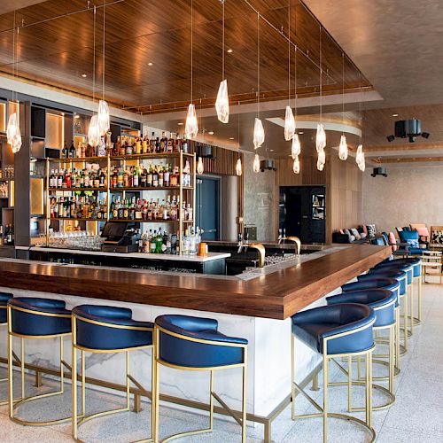 A modern bar with a fully stocked shelf, stylish bar stools, hanging lights, and a cozy seating area in the background.