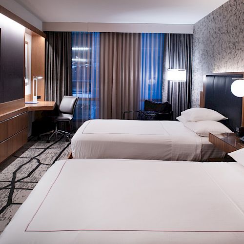 This image shows a modern hotel room with two neatly made beds, a desk, a chair, a flat-screen TV, and minimalistic decor.