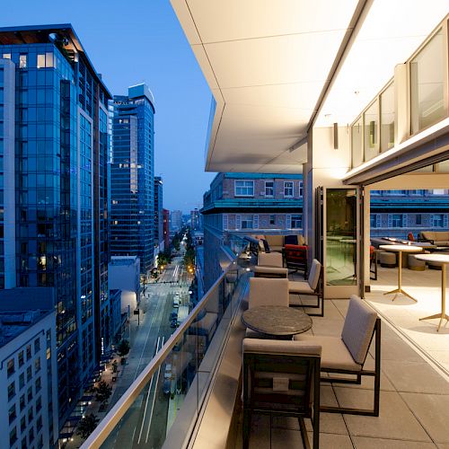 The image shows a modern cityscape at dusk with high-rise buildings and a sleek balcony featuring outdoor seating and tables, overlooking a bustling street.