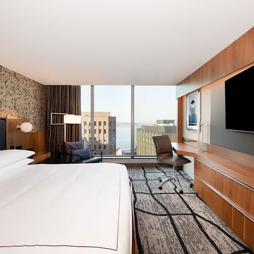 This image shows a modern hotel room with a large bed, a desk with a chair, a patterned carpet, a TV on the wall, and a view of buildings from the window.
