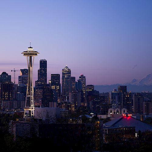 A twilight view of a city skyline featuring a prominent tower and distant mountain, with buildings illuminated as darkness falls, ending the sentence.