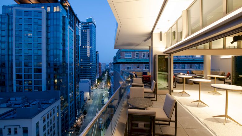 The image shows a modern outdoor balcony with seating and tables, overlooking a cityscape with tall buildings at dusk.