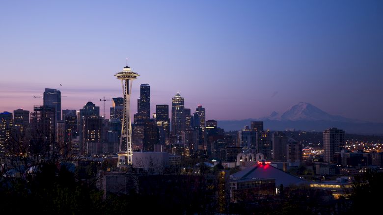 The image depicts the Seattle skyline at dusk, featuring the Space Needle prominently with city buildings and Mount Rainier visible in the background.